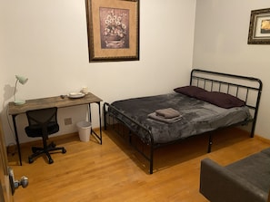 Ein Schlafzimmer mit Bett, Schreibtisch, Stuhl, Mülleimer und einem gerahmten Bild an der Wand.