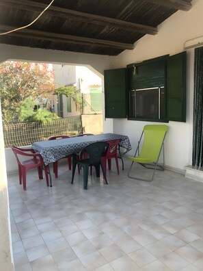 A covered patio with a table and chairs, a window with green shutters, and a view of a garden. - Holiday Home 'Marzamemi Home' with Private Terrace, Garden and Air Conditioning (Noto)