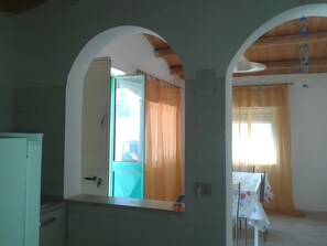 A room with a kitchen area, a dining table with a tablecloth, and a window with curtains. - Holiday Home 'Marzamemi Home' with Private Terrace, Garden and Air Conditioning (Noto)