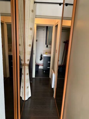 Un passadís estret amb terra de fusta, un cortineta blanca i un mirall que reflecteix una cuina amb armaris i una pica.