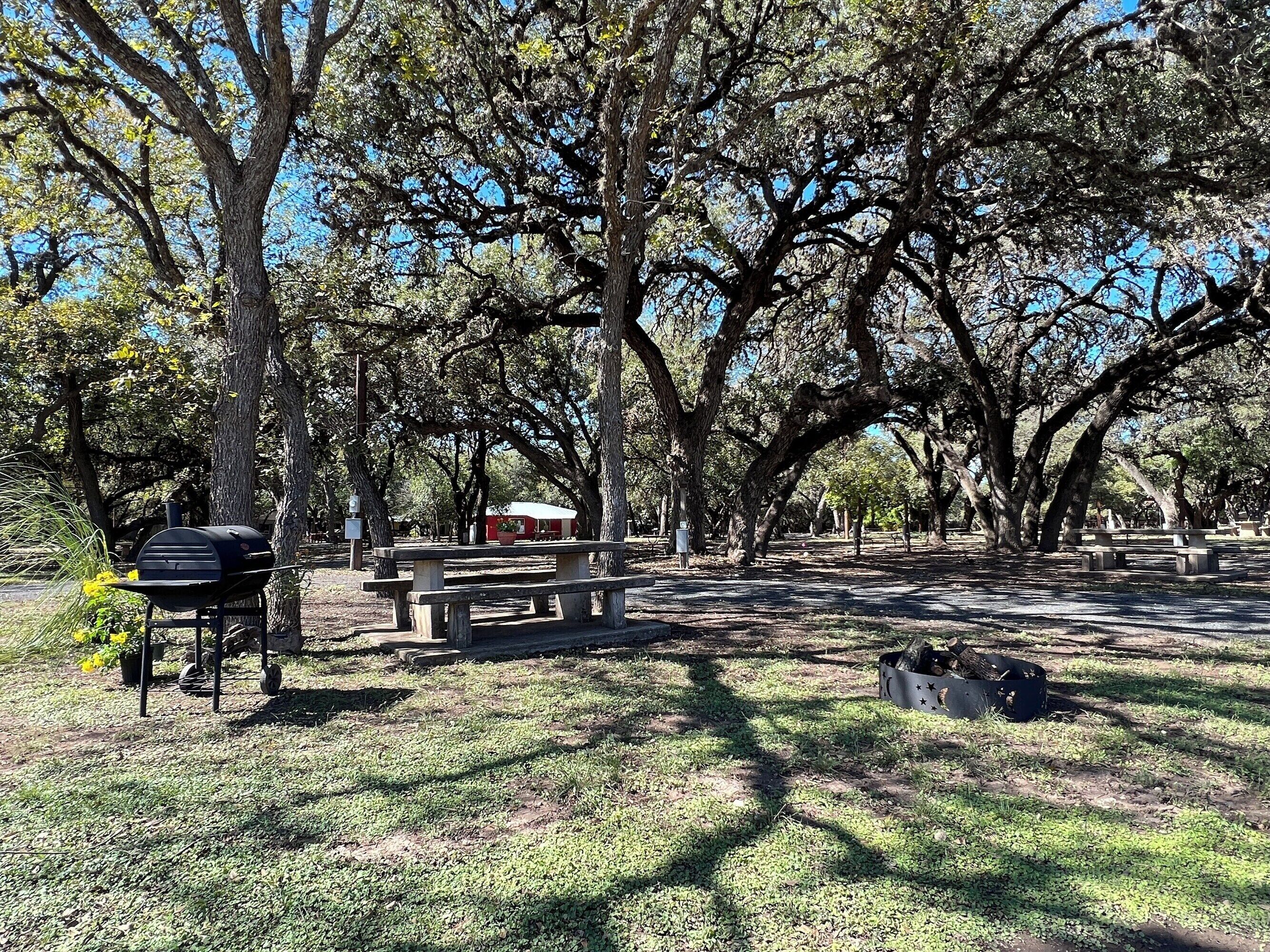 A park with picnic tables, a barbecue grill, and a red building.