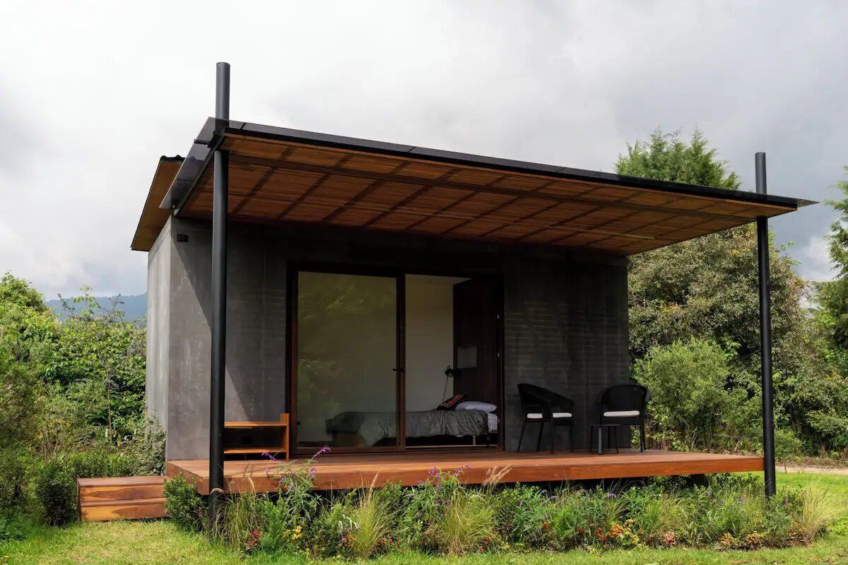 A modern, single-story building with a wooden deck, a sliding glass door leading to a bedroom, and two chairs outside.