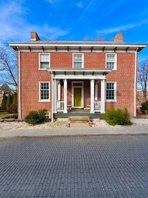A two-story brick house with a front porch, a yellow door, and a brick driveway.