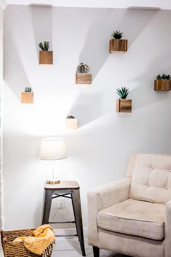 A room with a white wall, a beige armchair, a small wooden side table, a lamp, and several potted plants.