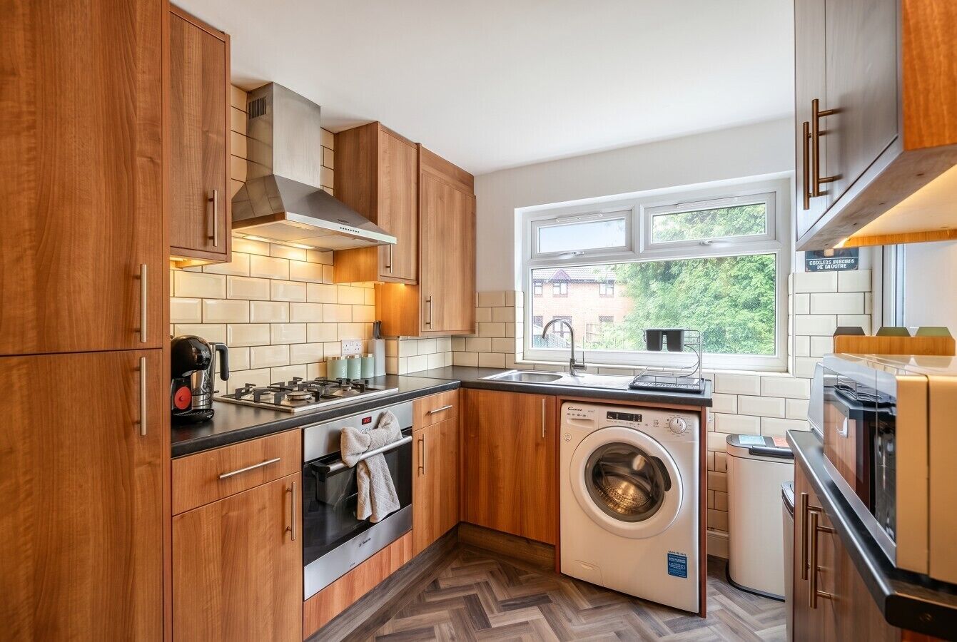 A compact kitchen with wooden cabinets, a built-in oven, a washing machine, and a window providing natural light.