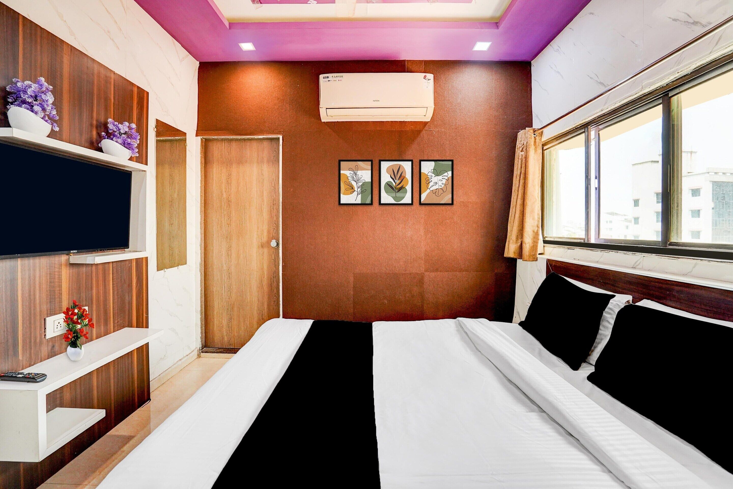 A hotel room with a bed, a TV, a wall-mounted air conditioner, and a window with a view of buildings.