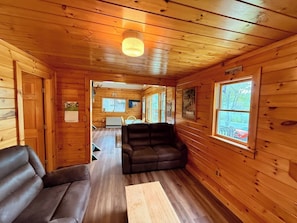 A cabin interior with wooden walls, a sofa, a table, and a window with a view. - Camp Up North | Water Access | Snowmobile & ATV (Rockwood)