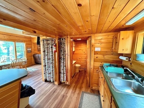 A wooden cabin interior with a kitchen, dining area, and a bathroom. - Camp Up North | Water Access | Snowmobile & ATV (Rockwood)