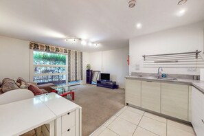 A modern living room with a sofa, a glass coffee table, and a television. There is a kitchen area with a sink and cabinets. - Bright 1BR Flat Near Canary Wharf & Stratford (London)