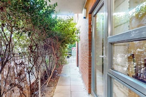 A narrow pathway flanked by dense foliage and a brick building with large windows. - Bright 1BR Flat Near Canary Wharf & Stratford (London)