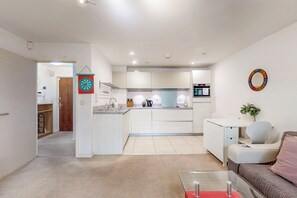 A modern kitchen with white cabinets, a central island, and a microwave. There is a dining area with a white table and a sofa. - Bright 1BR Flat Near Canary Wharf & Stratford (London)