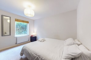 Ein Schlafzimmer mit einem Bett, einem Nachttisch, einem Spiegel, einem Fenster mit Blick auf Bäume und einer Deckenleuchte.