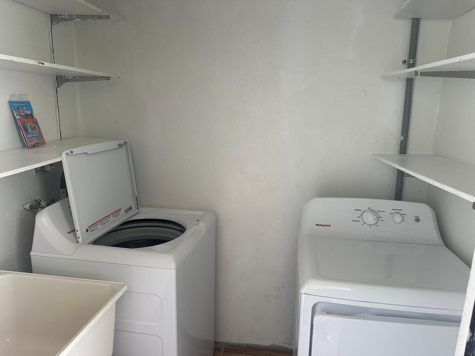 Une buanderie équipée d’un lave-linge et d’un sèche-linge à chargement frontal, d’une étagère sur laquelle se trouve une bouteille de lessive, et d’une étagère fixée au mur.