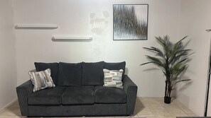 A dark grey sofa with two decorative pillows, a tall potted plant, and a framed abstract painting on the wall. - Cozy Nassau Hideaway – Local Island Charm (Nassau)