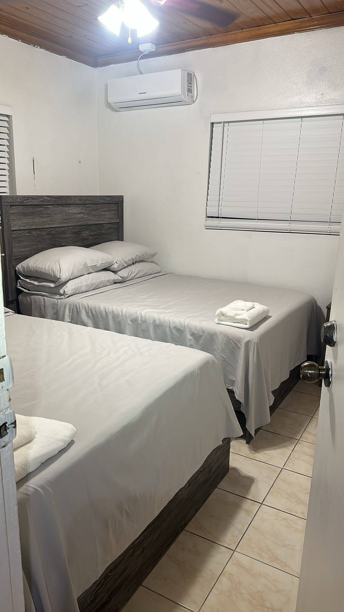 A bedroom with two beds, a window with blinds, an air conditioning unit, and a door with a handle.