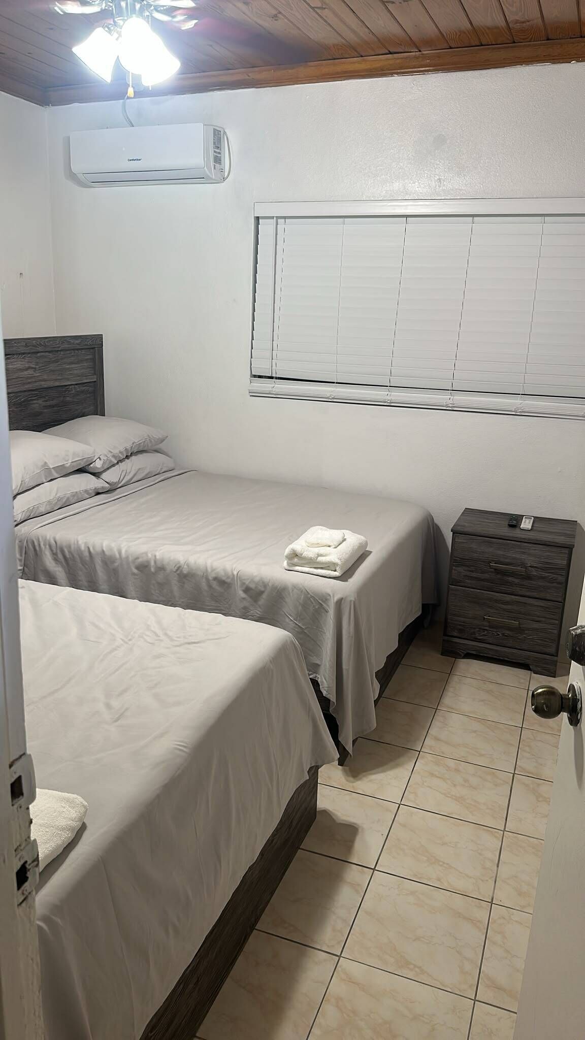 Un dormitorio con dos camas, una ventana con persianas, un aire acondicionado y un aparador de madera.