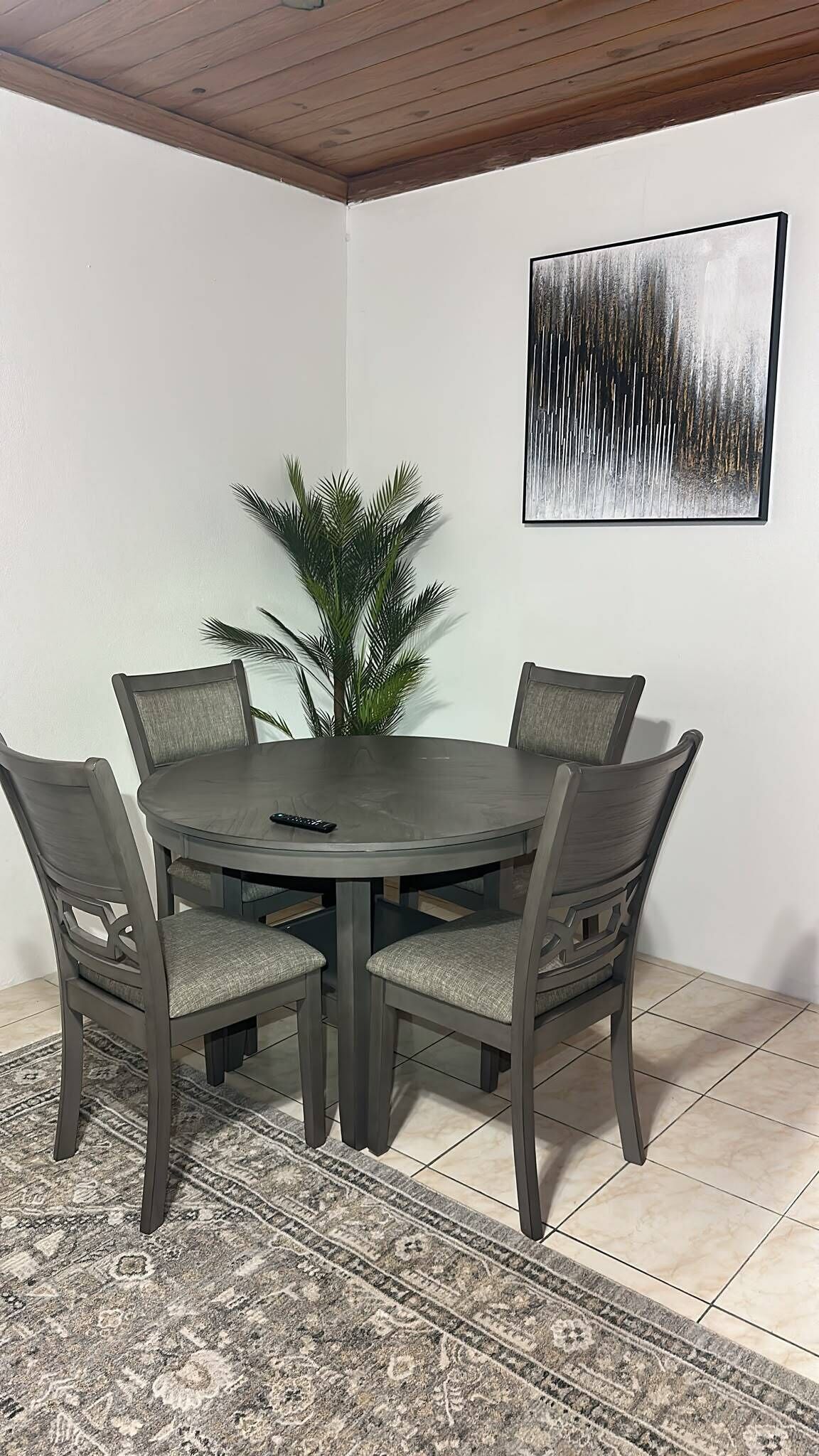 A dining area with a round table, four chairs, a remote control, a potted plant, and a framed abstract painting on the wall.