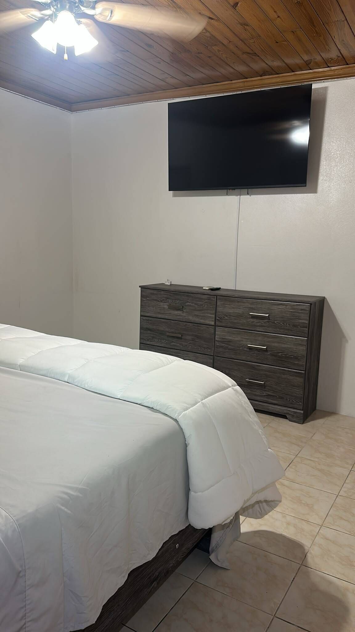 A bedroom with a bed, a dresser, a TV mounted on the wall, and a ceiling fan.
