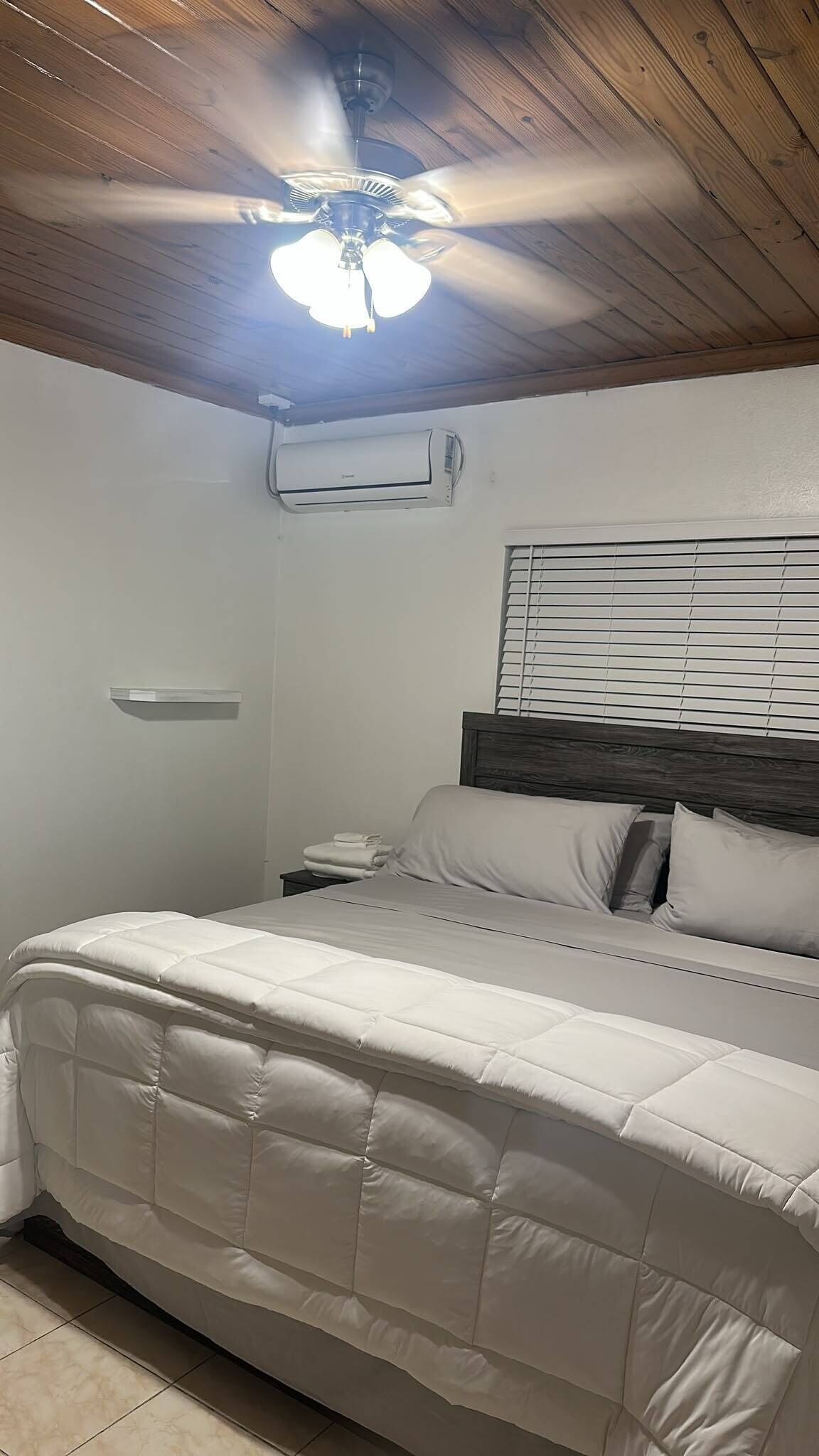 Ein Schlafzimmer mit einem Bett, Deckenventilator, Klimaanlage, Nachttisch mit Lampe und einem Fenster mit Jalousien.