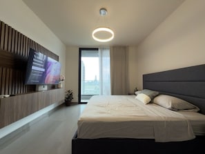 Quarto de hotel moderno com uma cama grande, televisão de tela plana na parede e uma janela ampla com cortinas.