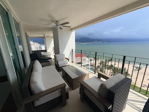 A balcony with a sofa, chairs, and a table, overlooking a beach and cityscape. - Luxury 3-Bedroom Apartment with Panoramic Ocean Views at the Grand Venetian (Puerto Vallarta)