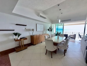 A modern dining area with a long table, white chairs, and a wooden cabinet. - Luxury 3-Bedroom Apartment with Panoramic Ocean Views at the Grand Venetian (Puerto Vallarta)