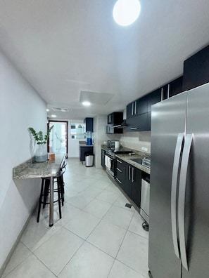 A modern kitchen with stainless steel appliances, a marble countertop, and a dining area with a bar stool. - Luxury 3-Bedroom Apartment with Panoramic Ocean Views at the Grand Venetian (Puerto Vallarta)