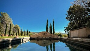 Una piscina tranquila con reflejos claros, rodeada de árboles bien recortados y una estructura de piedra.