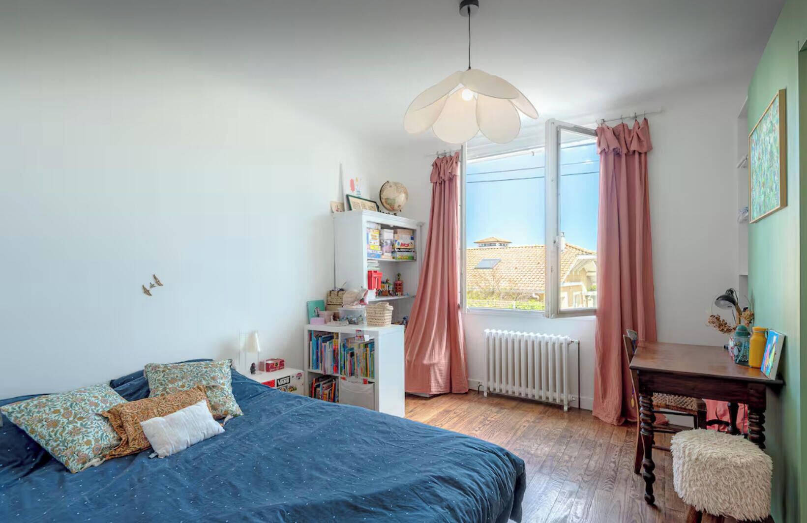 Un dormitorio con cama, escritorio, estantería, radiador y ventana con cortinas.