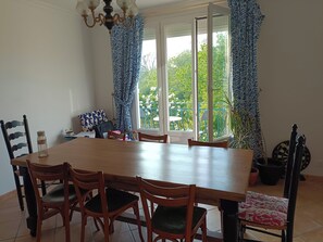 A dining area with a wooden table and chairs, a chandelier, and a view through large windows. - Quiet house 600 m from Morgat beach with beautiful wooded garden (Crozon)