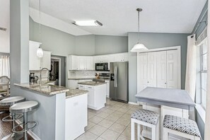 Una cocina moderna con armarios blancos, un refrigerador de acero inoxidable y un comedor con mesa y taburetes de bar.