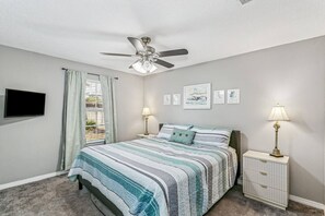 Una camera da letto con un letto, una ventola a soffitto, una TV, una finestra con tende, lampade da comodino e opere d'arte incorniciate al muro.