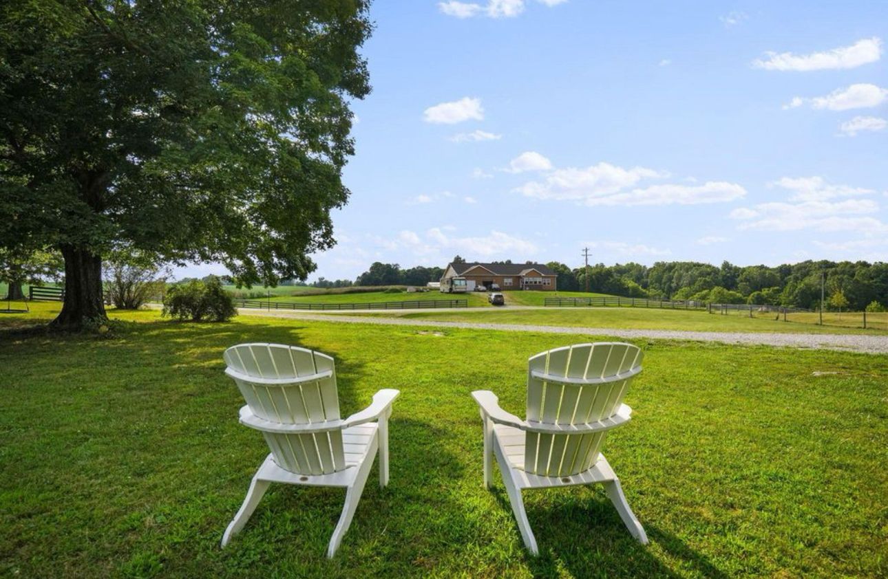 Een gazon met twee Adirondack-stoelen gericht op een huis met een grote boom links.