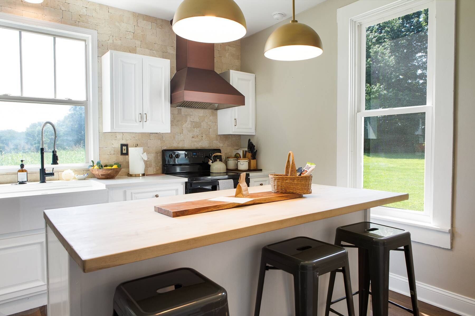 A kitchen with white cabinets, a large island, two bar stools, a hanging light fixture, and a window with a view of greenery.