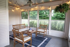 A covered porch with a wooden table and chairs, string lights, and large windows overlooking greenery.