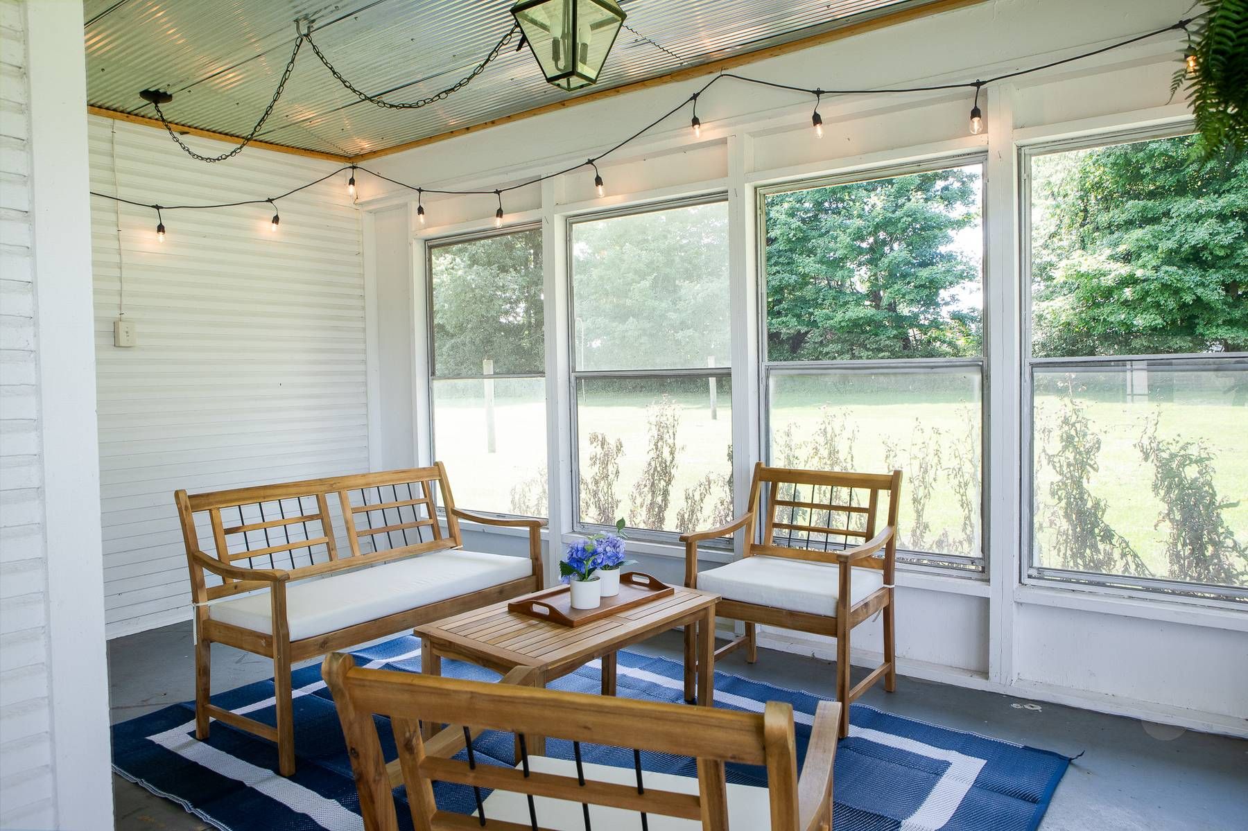 Un porche cubierto con muebles de madera, luces colgantes y grandes ventanales con vista a un jardín.