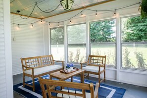 Un porche cubierto con muebles de madera, luces colgantes y grandes ventanales con vista a un jardín.