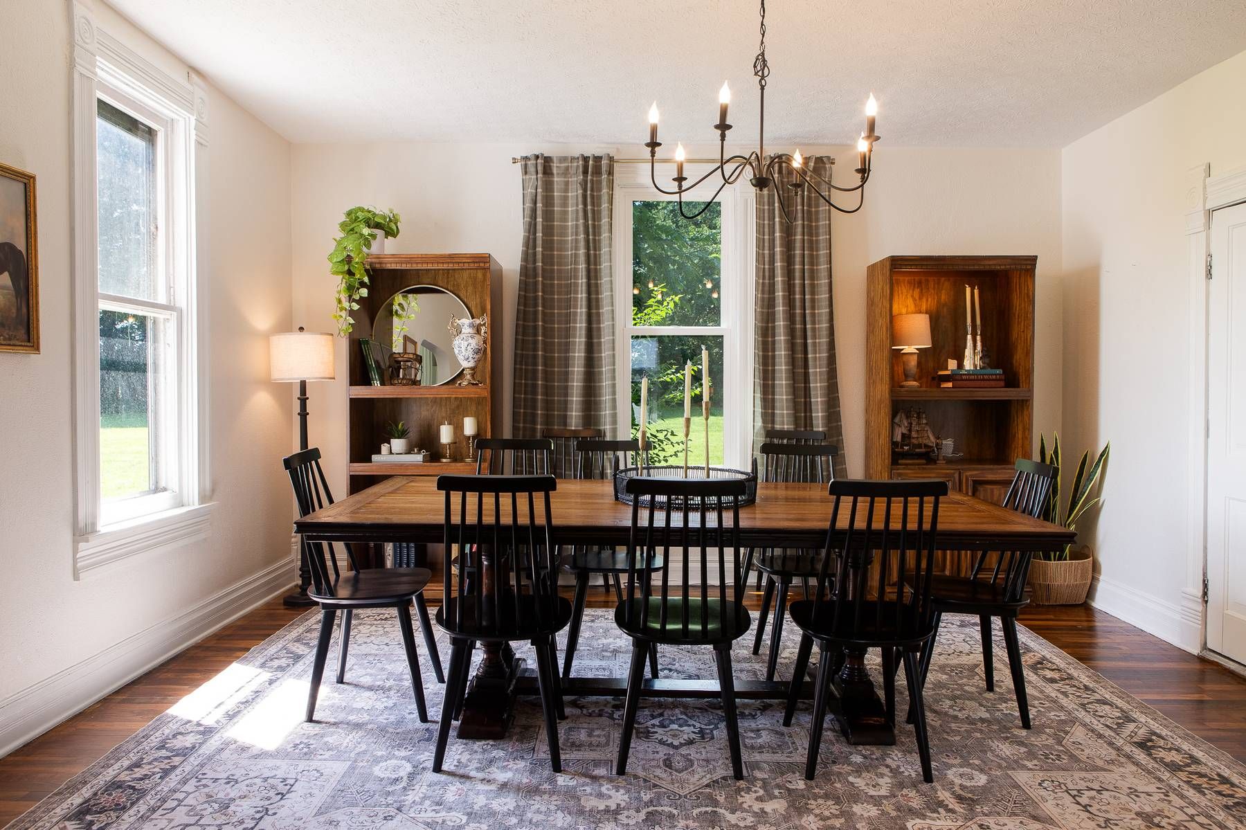 A dining room with a wooden table, black chairs, a chandelier, and a view of the outdoors through a window.