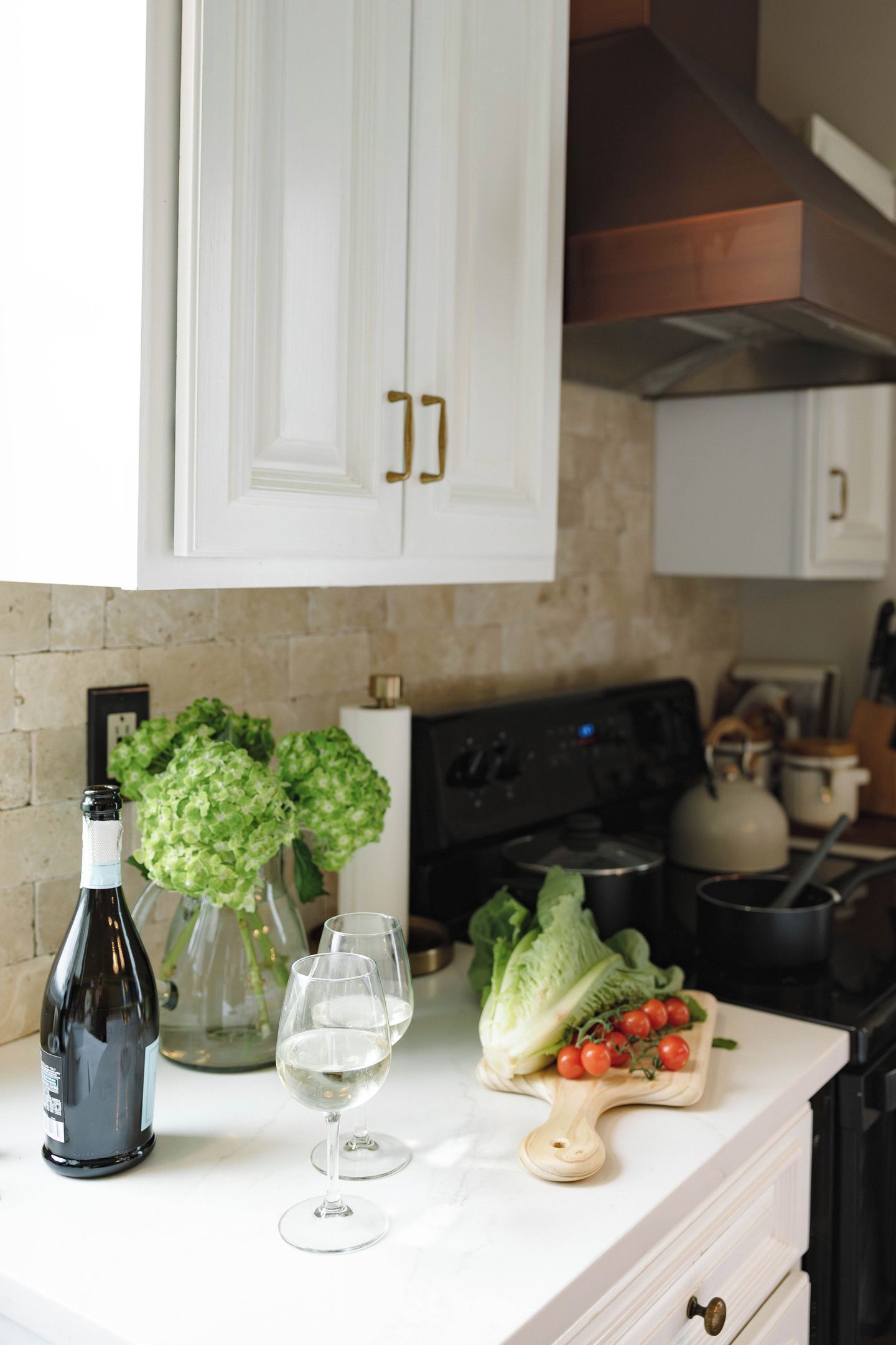 A kitchen counter with a wine bottle, glasses, a vase with flowers, and fresh vegetables.