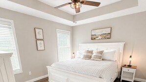 Un dormitorio con cama blanca, suelo de madera, ventilador de techo y mesita de noche con lámpara.