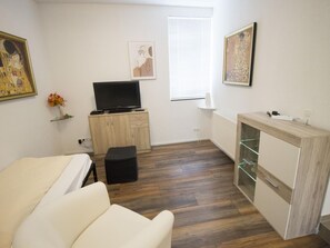 Ein Zimmer mit einem Bett, einem Fernseher, einer kleinen Kommode und einem Fenster mit Jalousien.