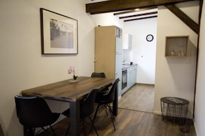 A dining area with a wooden table, black chairs, a framed picture on the wall, a kitchen in the background, and a small round table with a wireframe base. - NEW! Apartment Beamed room (Alsfeld)