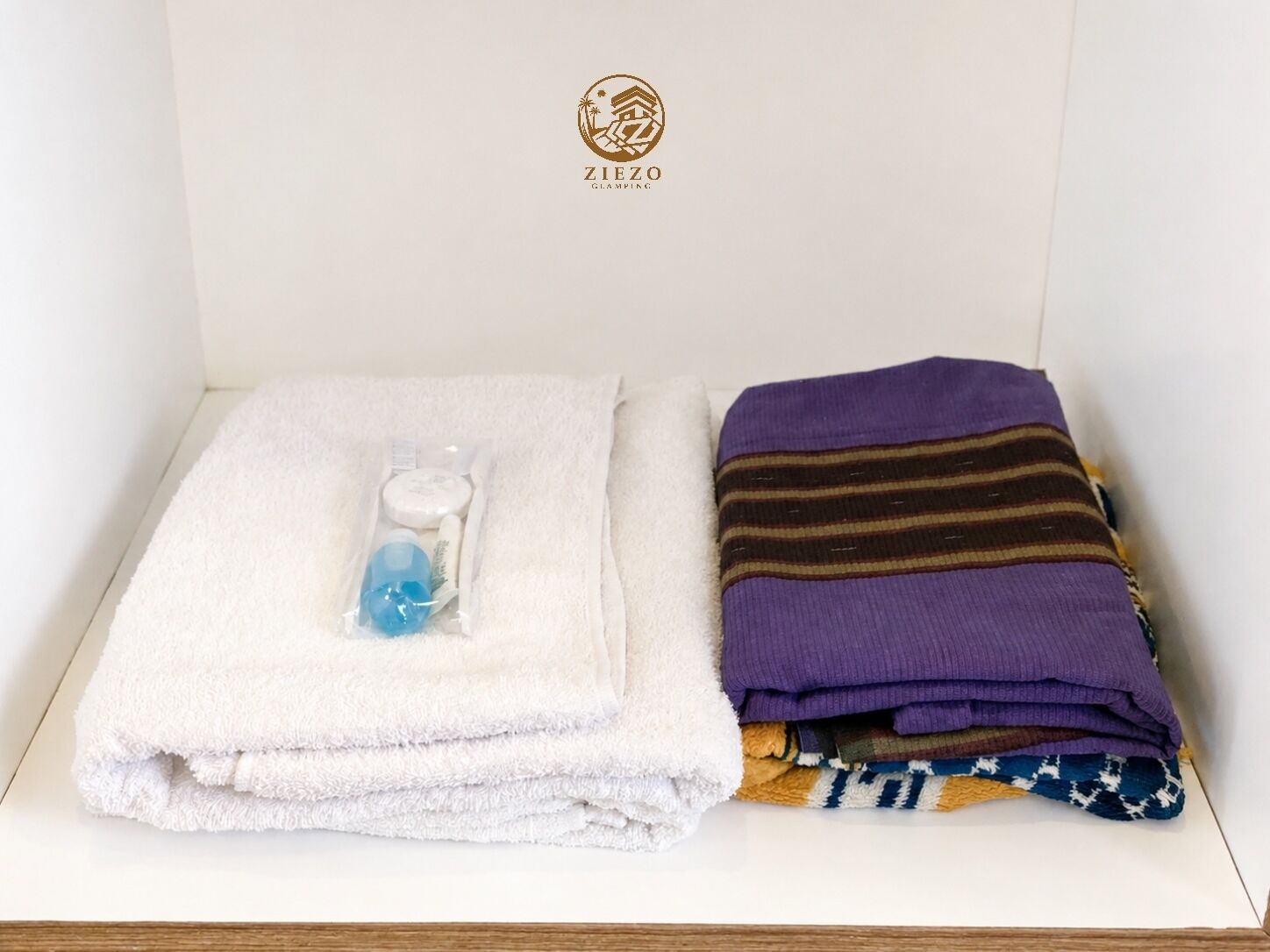 Une serviette blanche soigneusement pliée, une serviette violette et une pile de vêtements pliés dans une boîte blanche ornée d’un logo.