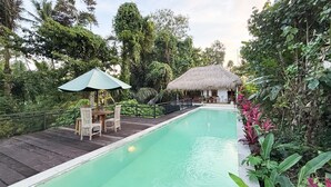 Ein Pool mit Holzterrasse, einer Reetdachkonstruktion und üppiger Bepflanzung.