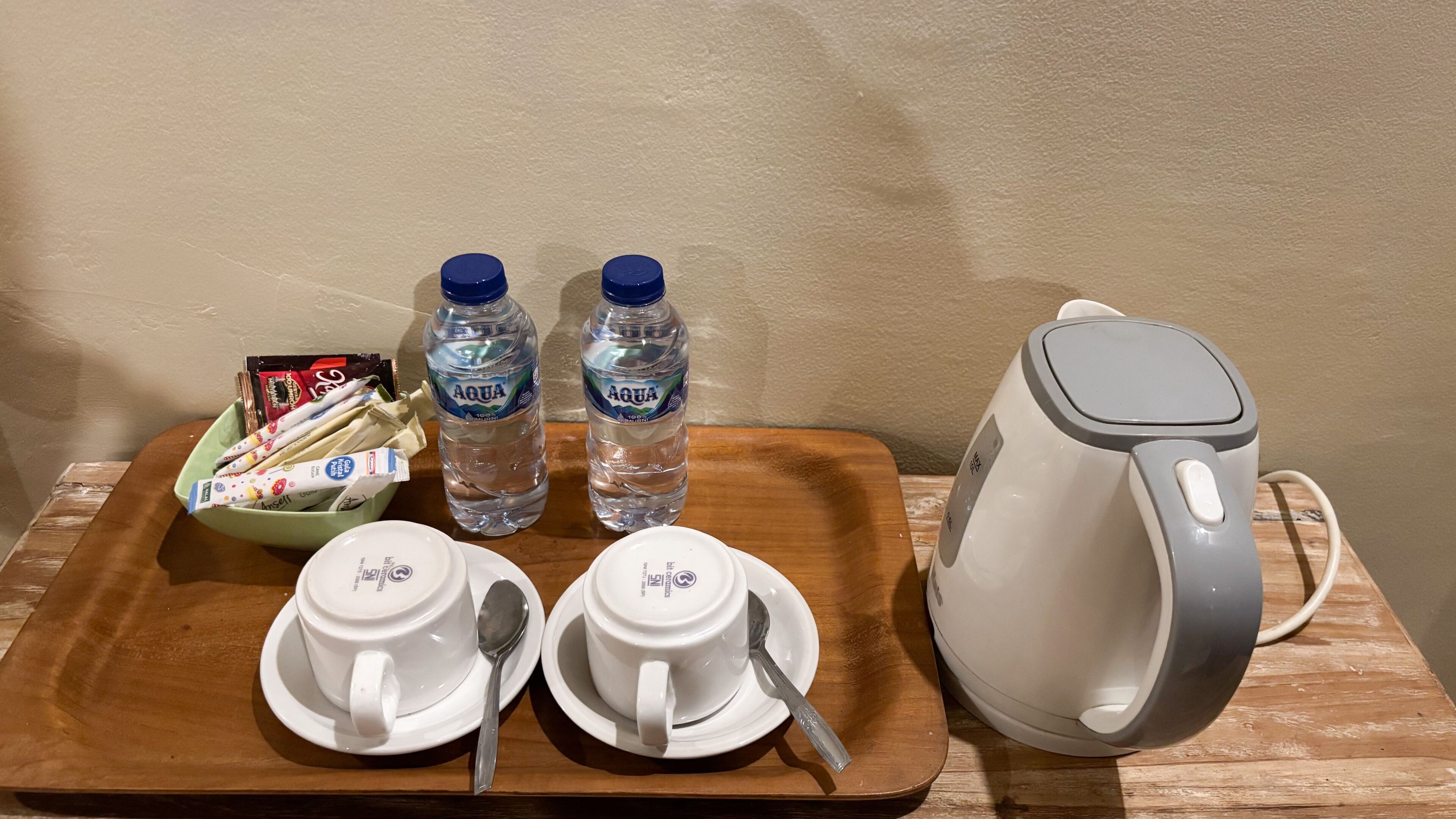 A tray with two white cups, two spoons, two bottles of water, a bowl of snacks, and a kettle.
