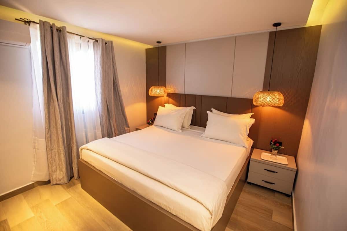 Una cama bien hecha con sábanas blancas, una mesita de noche con lámpara y un cabecero de madera.
