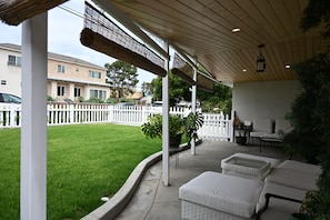 Ein überdachter Patio-Bereich mit Gartenmöbeln, einem weißen Zaun und einem Rasen.