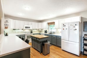 A modern kitchen with white cabinets, a central island, stainless steel appliances, and a view of greenery outside.