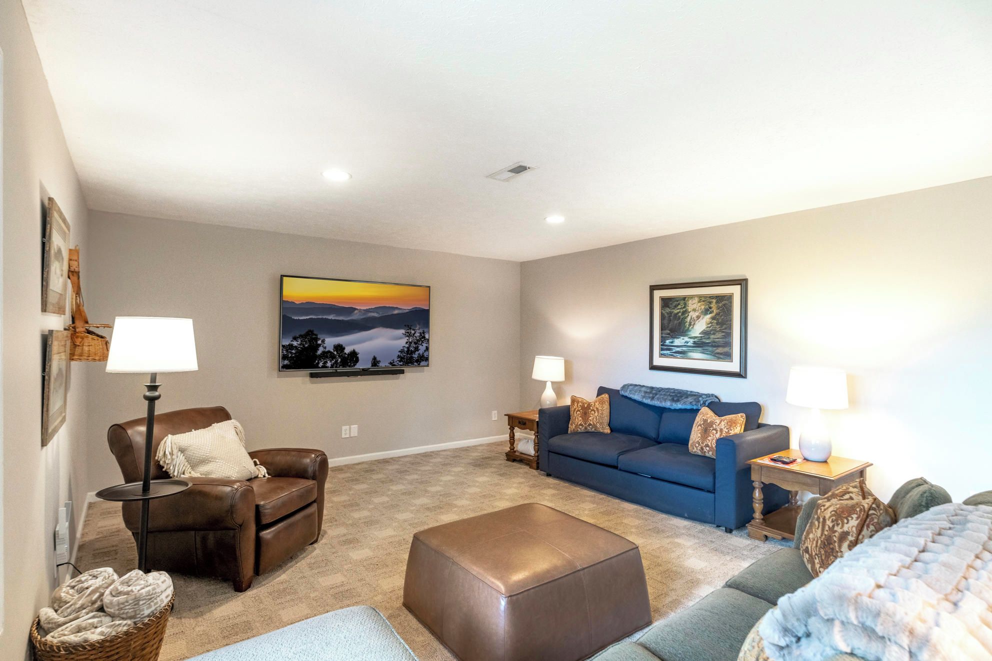 A spacious living room with a brown leather sofa, a blue sofa, a brown leather ottoman, and a grey armchair. There are two wall-mounted lamps and two framed pictures on the walls.