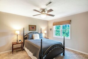 A bedroom with a four-poster bed, two bedside tables with lamps, a ceiling fan, a window with a view of trees, and a wall-mounted clock.